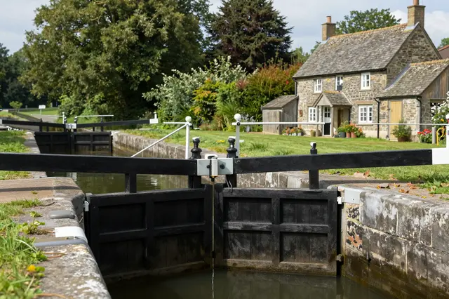 Dark wooden canal lock gates sit beside a stone lockkeeper's cottage on a sunny summer day