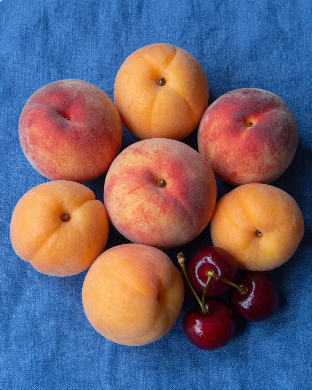 Top-down view of ripe peaches, apricots, and cherries arranged on a textured blue linen cloth.