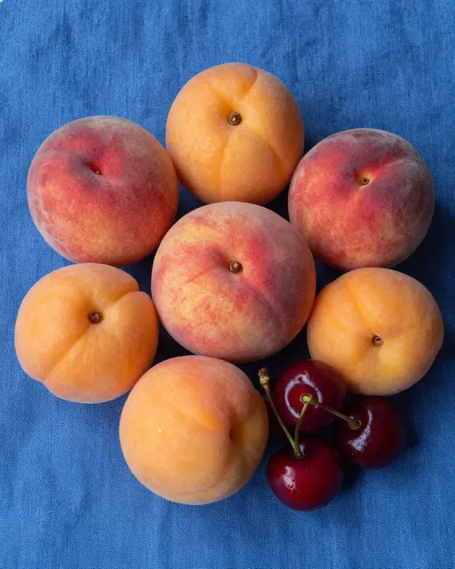 Top-down view of ripe peaches, apricots, and cherries arranged on a textured blue linen cloth.