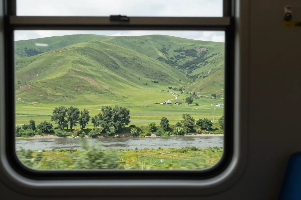 Lush green hills and a river are seen through a moving train window on a bright summer day