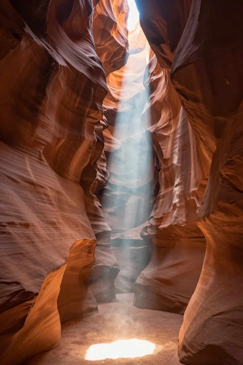 A narrow shaft of sunlight falls through a slot canyon opening onto the sandy canyon floor between flowing sandstone walls