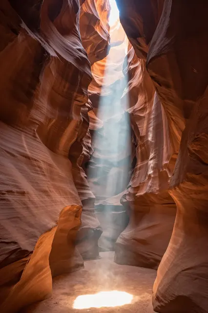 A narrow shaft of sunlight falls through a slot canyon opening onto the sandy canyon floor between flowing sandstone walls
