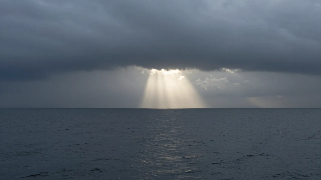 A narrow shaft of sunlight breaks through dark storm clouds to fall across a calm dark ocean