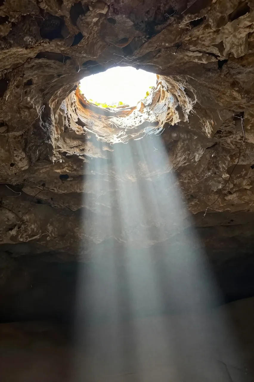 Bright sunbeams stream down through a circular opening in a rough cave ceiling