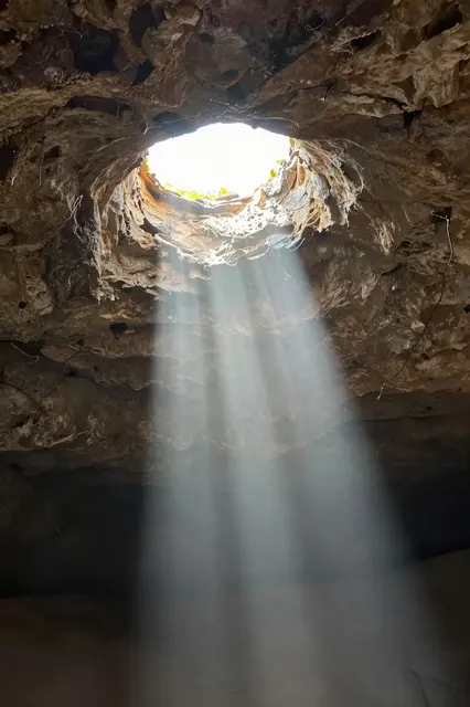 Bright sunbeams stream down through a circular opening in a rough cave ceiling