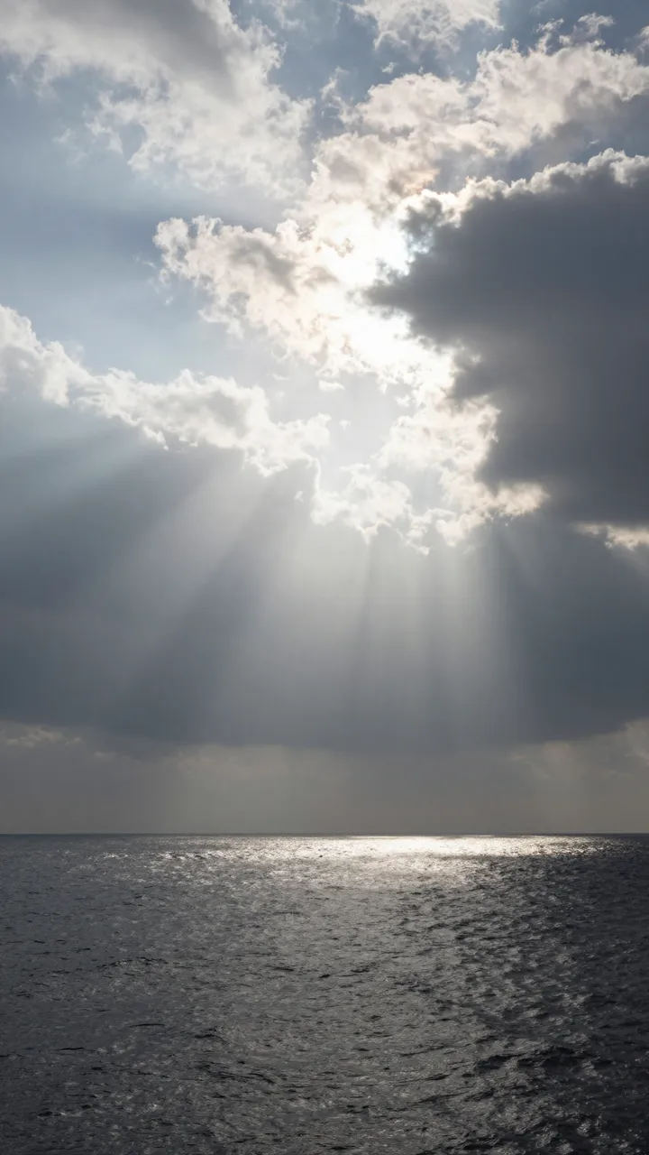 Sunbeams stream through broken clouds down to a glistening rippled sea surface