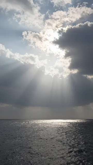 Sunbeams stream through broken clouds down to a glistening rippled sea surface
