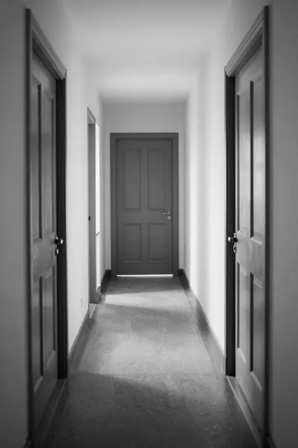 Black and white view down a narrow sunlit residential hallway lined with paneled wooden doors