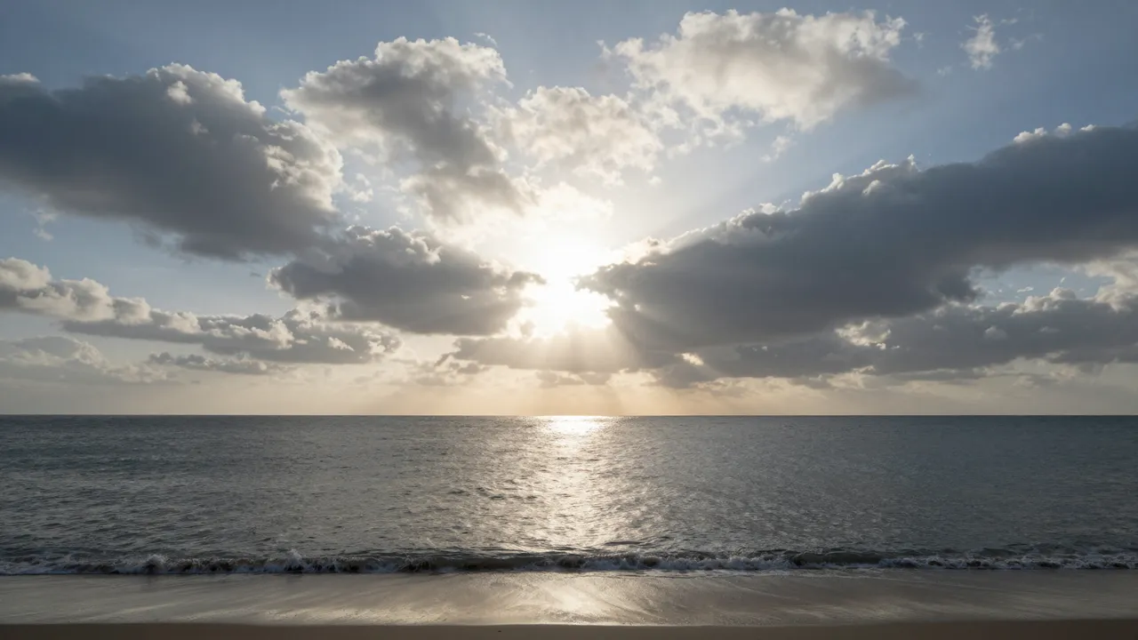 Bright sun peeking through scattered clouds over a calm sea and sandy beach