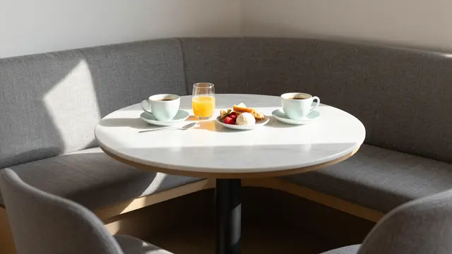 Round marble breakfast table with coffee, orange juice and pastries sits in a sunlit upholstered corner banquette.