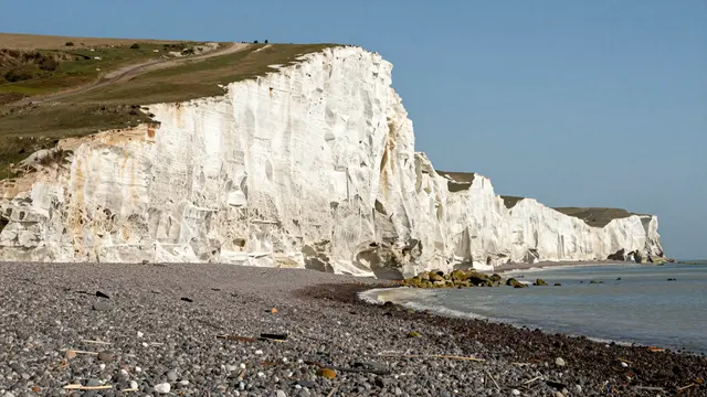 Bright white chalk cliffs rise above a pebbled shore under a clear blue sky on a sunny day