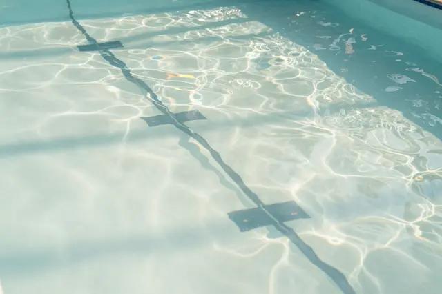 Rippled sunlight patterns cover the pale bottom of an empty outdoor swimming pool with a lane line