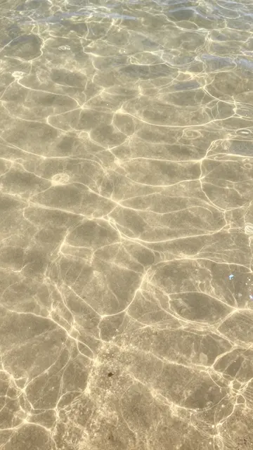 Top-down view of clear shallow water over rippled sand with glowing sun refraction patterns.