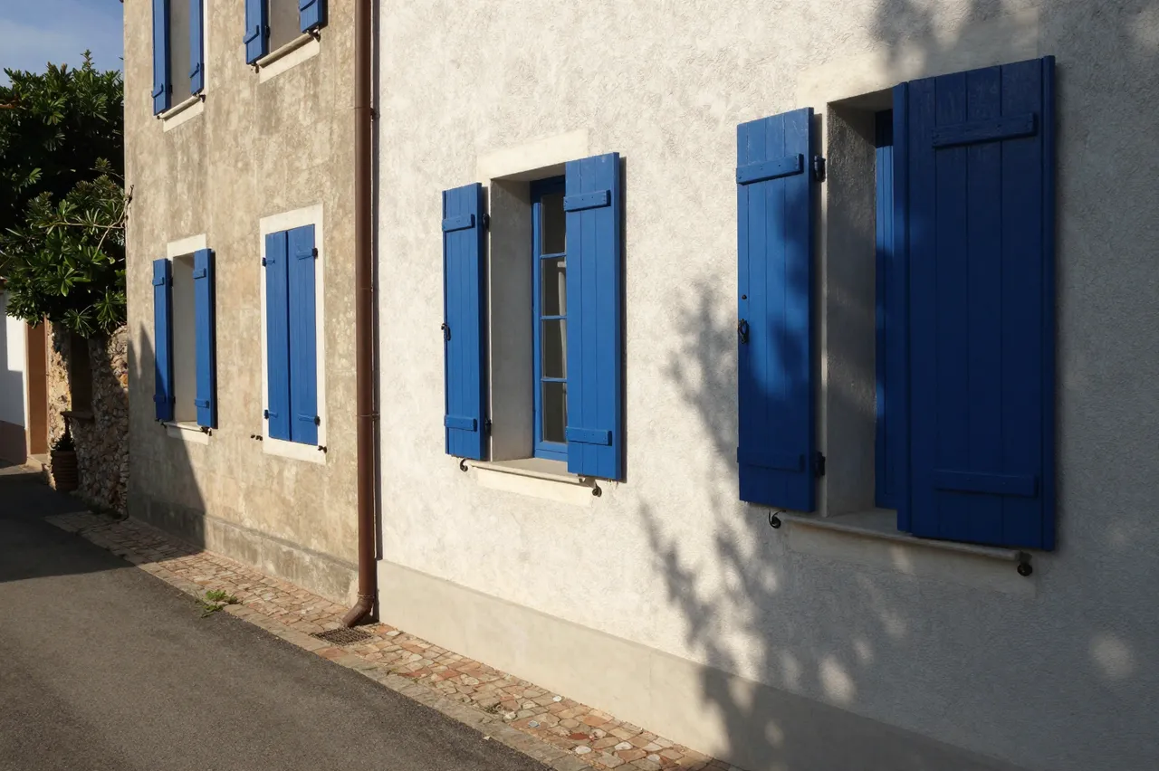 Sunlit plaster village building with bright blue wooden shutters along a quiet paved lane, cast with deep afternoon shadows