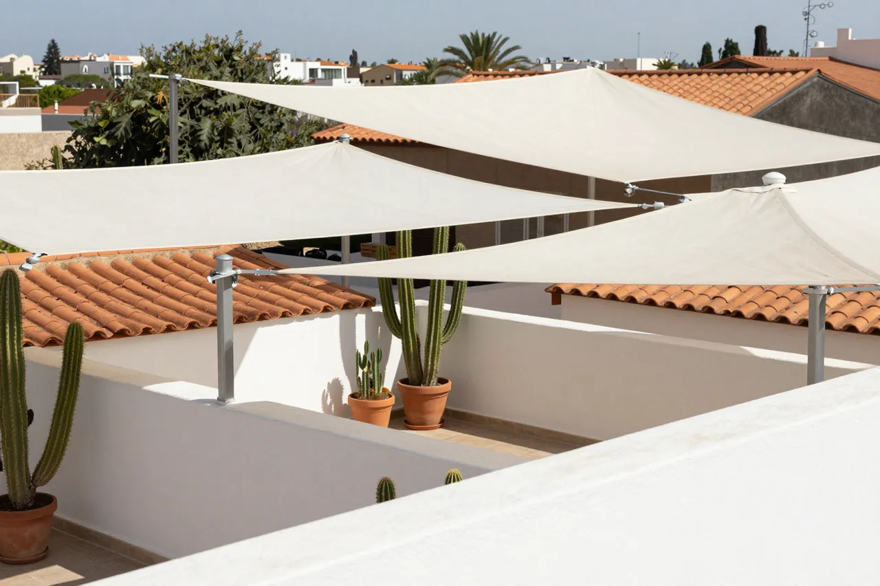 Midday view of a rooftop terrace shaded by white sails with potted cacti overlooking terracotta rooflines