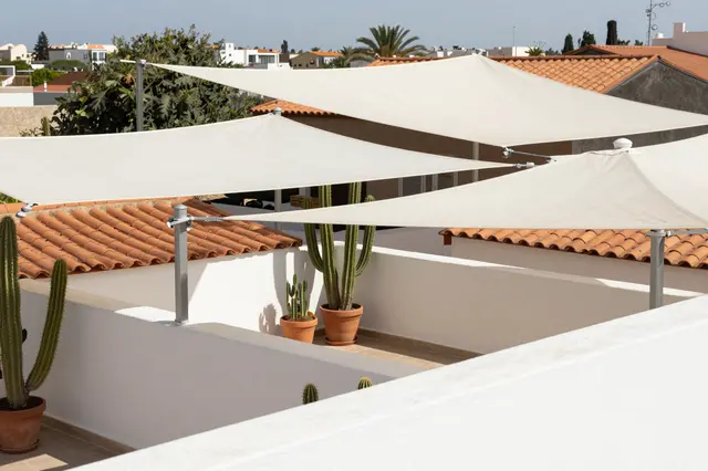 Midday view of a rooftop terrace shaded by white sails with potted cacti overlooking terracotta rooflines