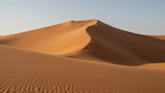 Tall orange sand dune with wind ripples, one face in shadow under a pale clear sky at sunrise