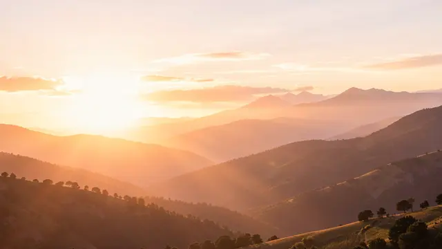 Warm soft sunrise light spills over layered hazy rolling hills with wide empty sky space