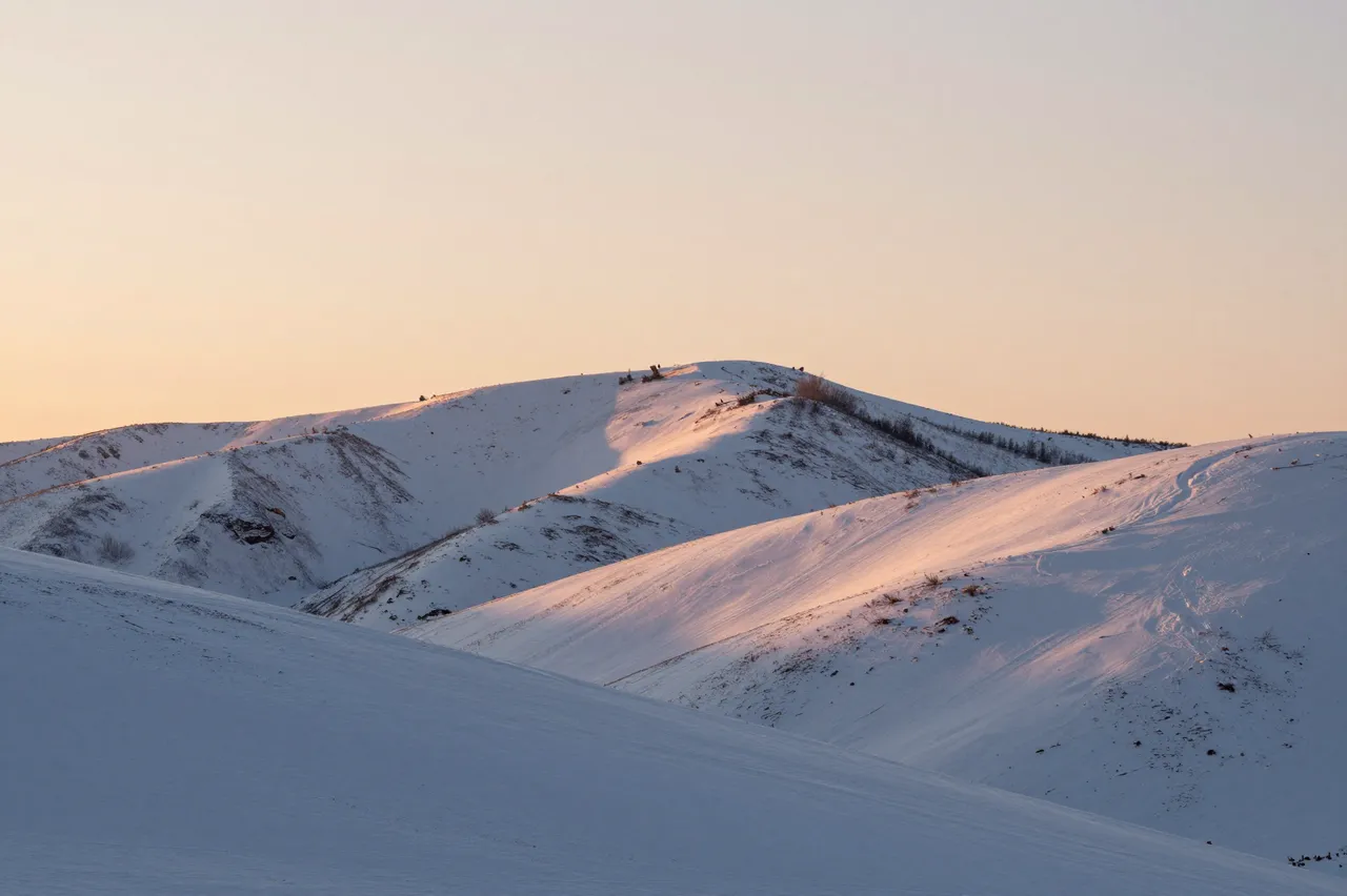 Soft peach sunrise light illuminates rolling snow-covered hills in a quiet winter landscape