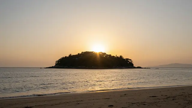 Golden sunset glows behind a forested small island viewed from a sandy ocean beach