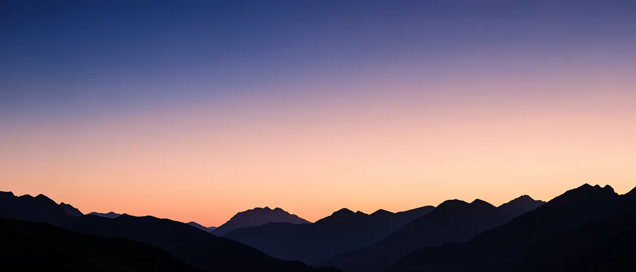 Smooth indigo to peach twilight sky above layered dark mountain silhouettes in an ultrawide frame