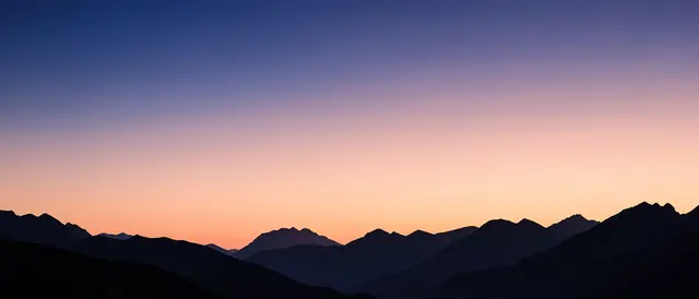 Smooth indigo to peach twilight sky above layered dark mountain silhouettes in an ultrawide frame