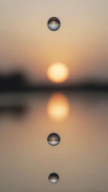 Vertical close-up of reflective droplets arranged over a soft gradient sunset backdrop with mirrored light