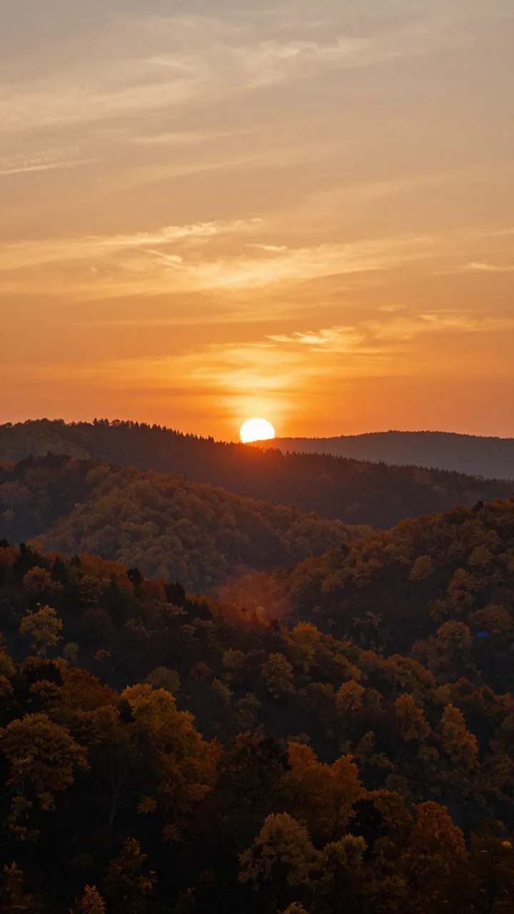 Bright orange sunset hangs low over layered forested autumn hills beneath a wispy warm sky