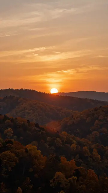 Bright orange sunset hangs low over layered forested autumn hills beneath a wispy warm sky