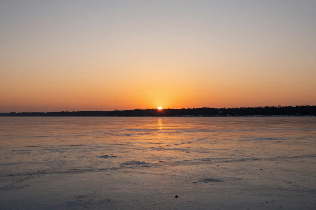Warm orange sunset light reflects across the smooth surface of a frozen lake with a distant treeline on the horizon
