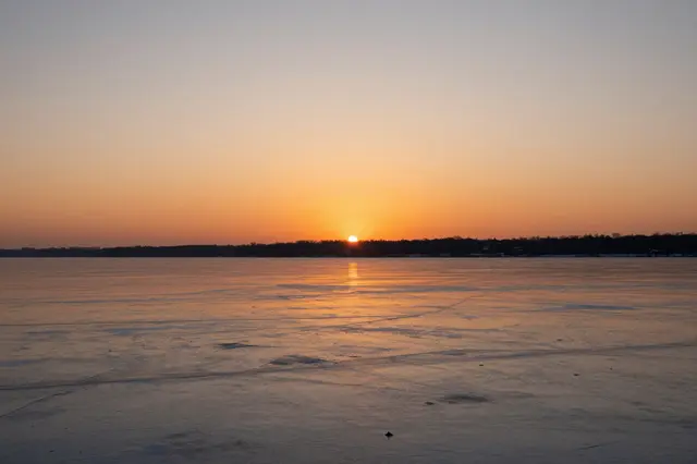 Warm orange sunset light reflects across the smooth surface of a frozen lake with a distant treeline on the horizon