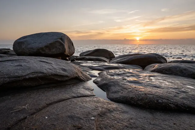 Golden sunset light illuminates smooth wet boulders along a coastal beach overlooking the ocean.