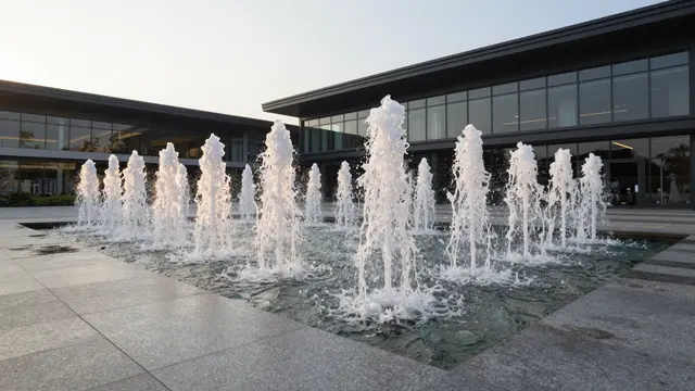 Row of vertical fountain jets rise from granite paving in front of a modern glass building at sunset