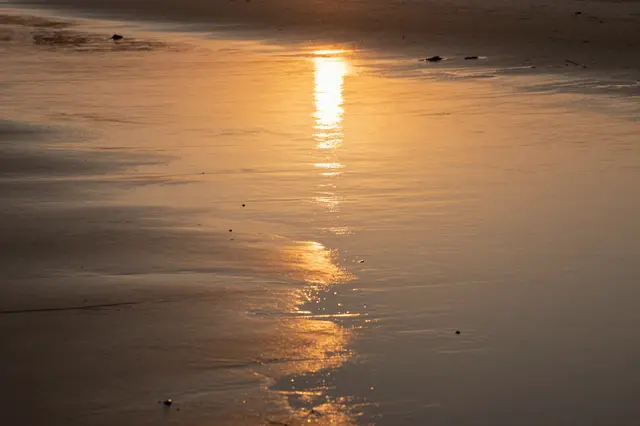 Wet low-tide beach sand reflects a bright orange golden sunset glow in a smooth vertical path.