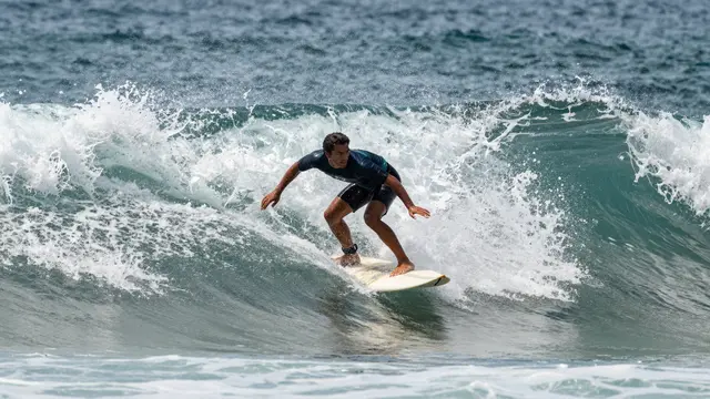 A male surfer crouches balancing on a white surfboard while riding a curling turquoise ocean wave