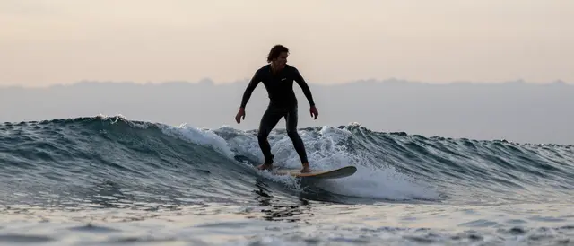 A surfer in a black wetsuit rides a small ocean surfboard during soft dusk light