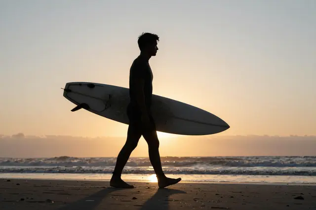 Silhouette of a surfer carrying a board while walking along a sandy beach at sunset