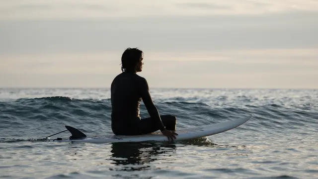 A silhouetted adult surfer sits on a surfboard in calm ocean waters at dawn