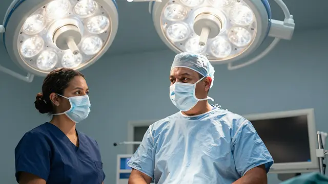 Two masked medical staff stand under operating lights in a hospital surgical suite