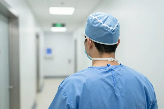 A surgeon in blue scrubs and cap stands facing down a bright hospital corridor, viewed from behind.