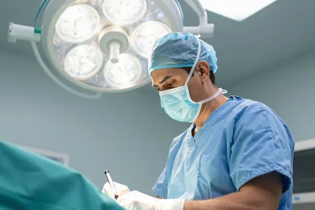 Masked male surgeon in blue scrubs concentrates under surgical lights next to teal drapes