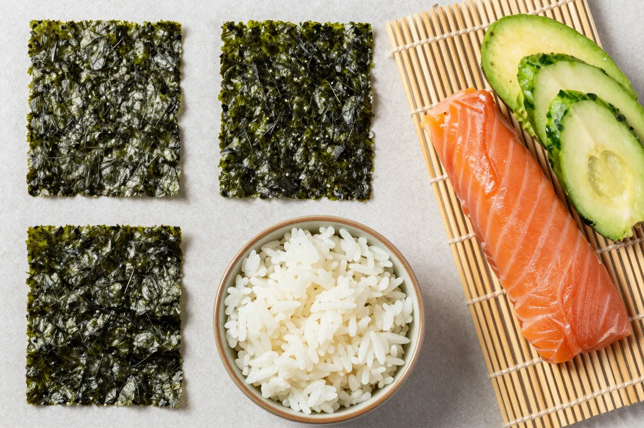 Top-down flat lay of sushi preparation ingredients arranged on a light neutral surface with a bamboo rolling mat.