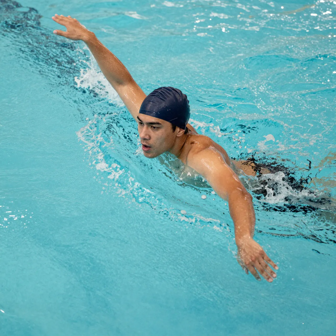 Young adult male swimmer with a dark swim cap performs a freestyle stroke in turquoise pool water viewed from above.