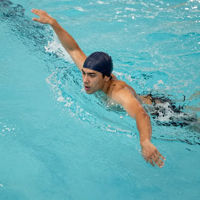 Young adult male swimmer with a dark swim cap performs a freestyle stroke in turquoise pool water viewed from above.