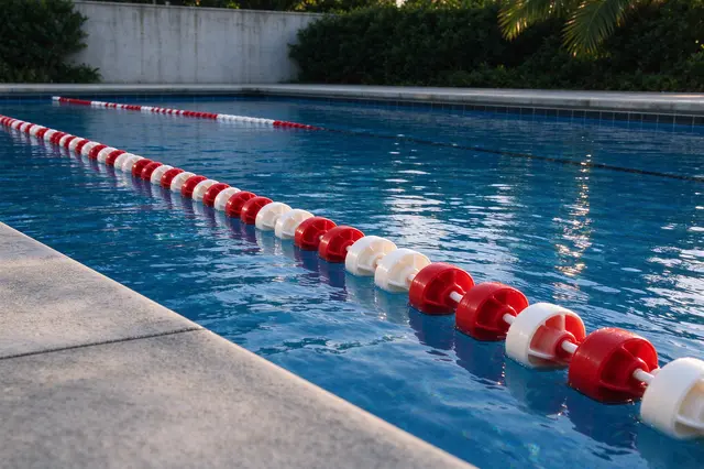 Red and white lane division floats stretch across a bright blue outdoor swimming pool from the concrete deck edge at water