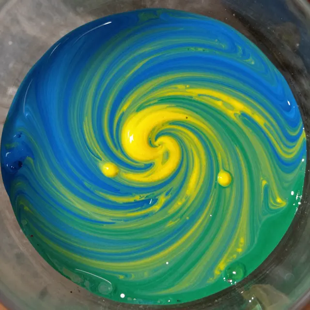 A close-up macro view of wet blue and yellow paint swirling into green on glass in a tight spiral pattern