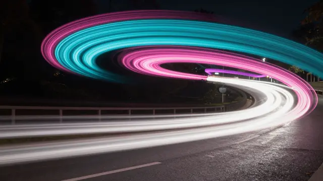 Long exposure captures swirling pink, cyan and white light trails arcing over a dark curved night road