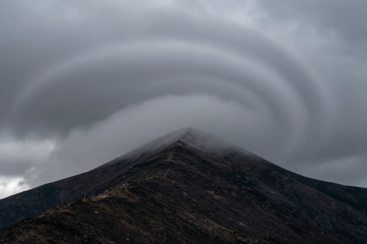 Layered swirling storm clouds wrap around the sharp rocky peak of a dark mountain summit.
