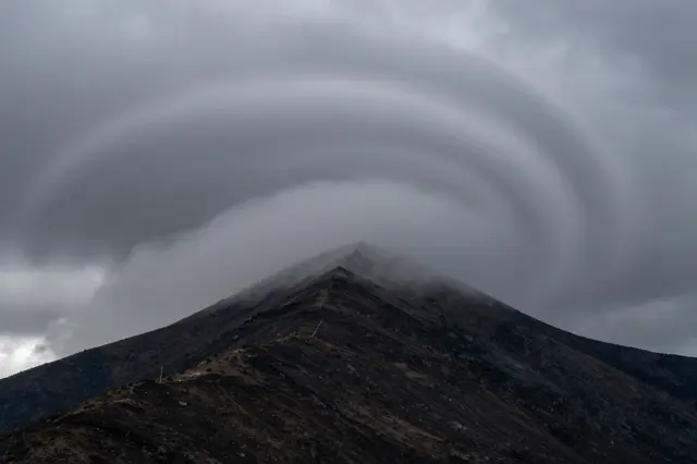 Layered swirling storm clouds wrap around the sharp rocky peak of a dark mountain summit.