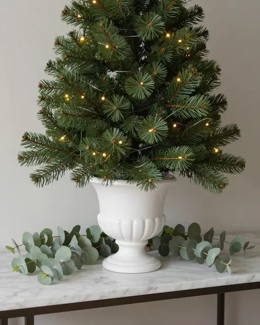 A small lit tabletop Christmas tree sits in a white ceramic urn on a marble console with eucalyptus sprigs.